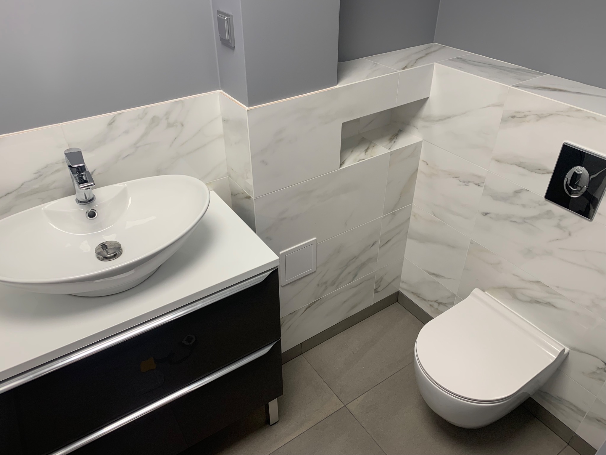 Nowoczesna łazienka z umywalką nablatową na czarnej szafce, wiszącą muszlą klozetową i marmurowymi płytkami na ścianach.