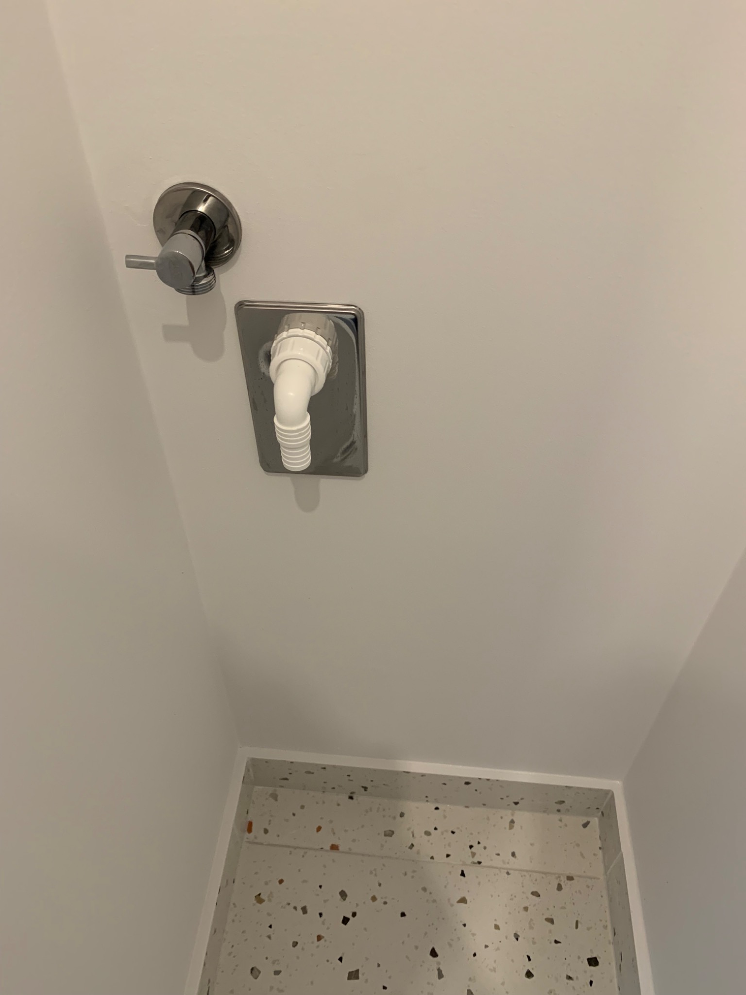 Chromowany zawór i odpływ umywalki zamontowane na białej ścianie, widoczny fragment umywalki z wzorem lastryko.