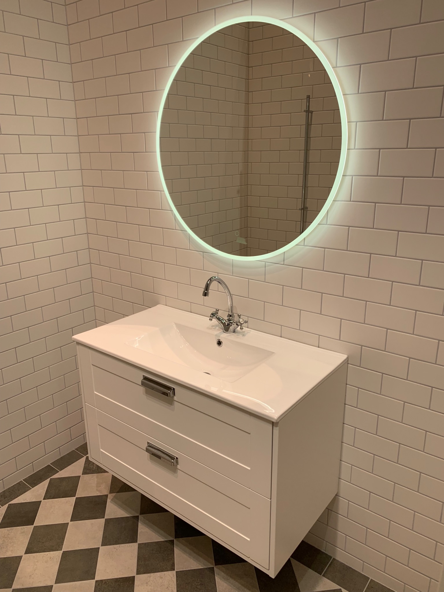 Wykończona łazienka z białą umywalką z szafką, podświetlanym okrągłym lustrem i szarą mozaiką na podłodze ułożoną w szachownicę.