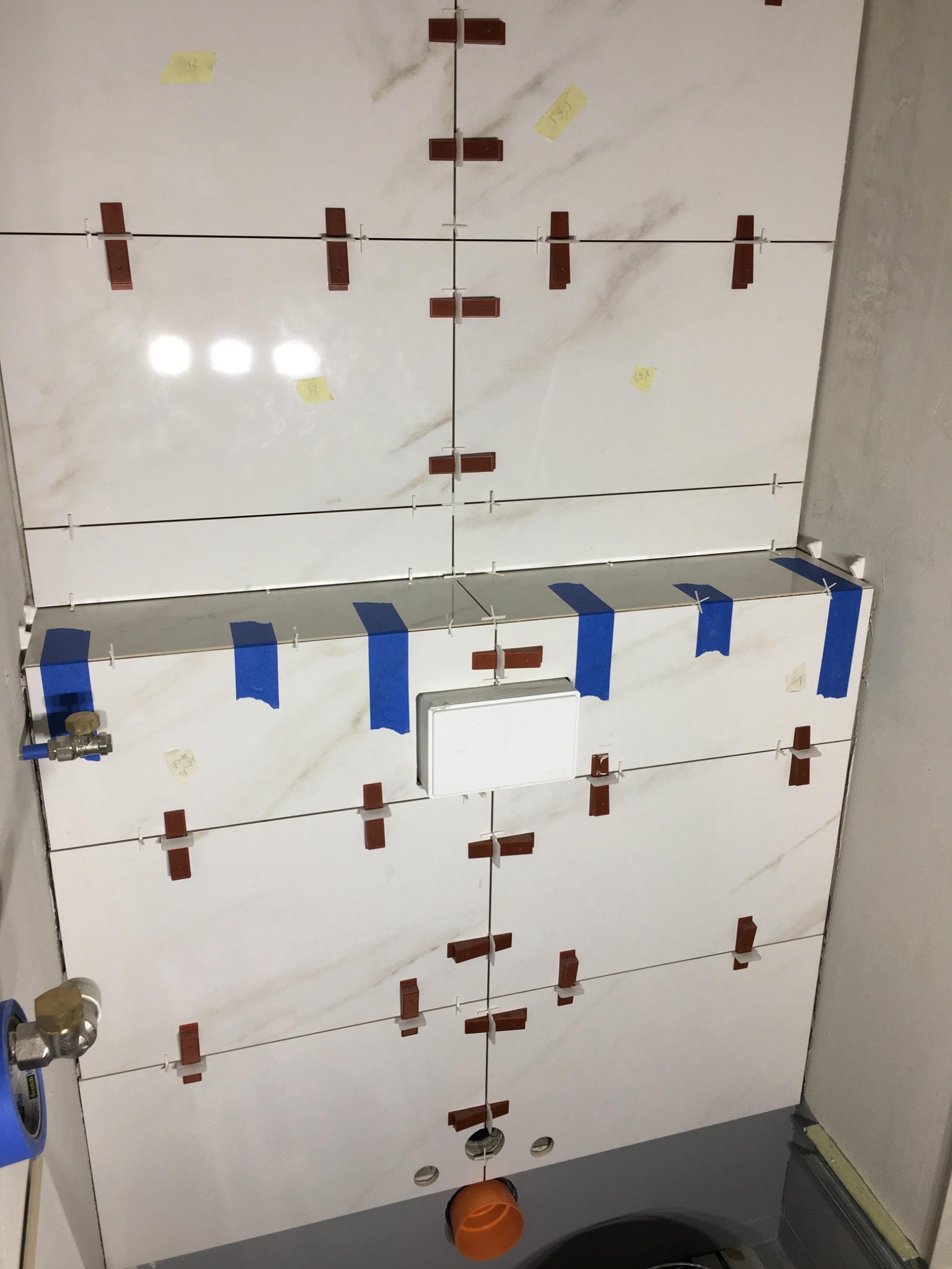 Montaż białych płytek imitujących marmur na ścianie w łazience, z użyciem krzyżyków dystansowych i taśmy maskującej, widoczne przyłącza wodne.