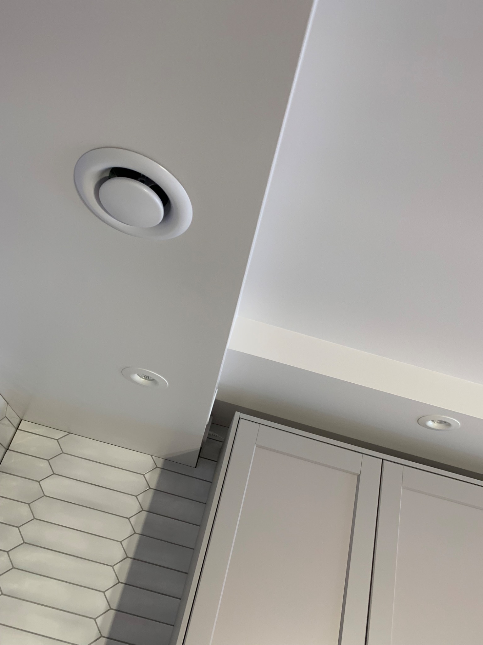 Fragment kuchni po remoncie: biały sufit z okrągłym nawiewnikiem i punktowym oświetleniem, płytki heksagonalne na ścianie, górne szafki kuchenne w jasnym odcieniu szarości.