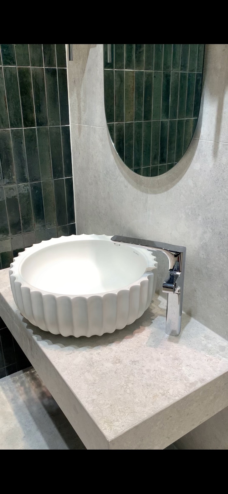 Nowoczesna łazienka z okrągłą, białą umywalką o żłobkowanej fakturze, osadzoną na szarym blacie z betonu architektonicznego, z chromowaną baterią i zielonymi płytkami w tle.