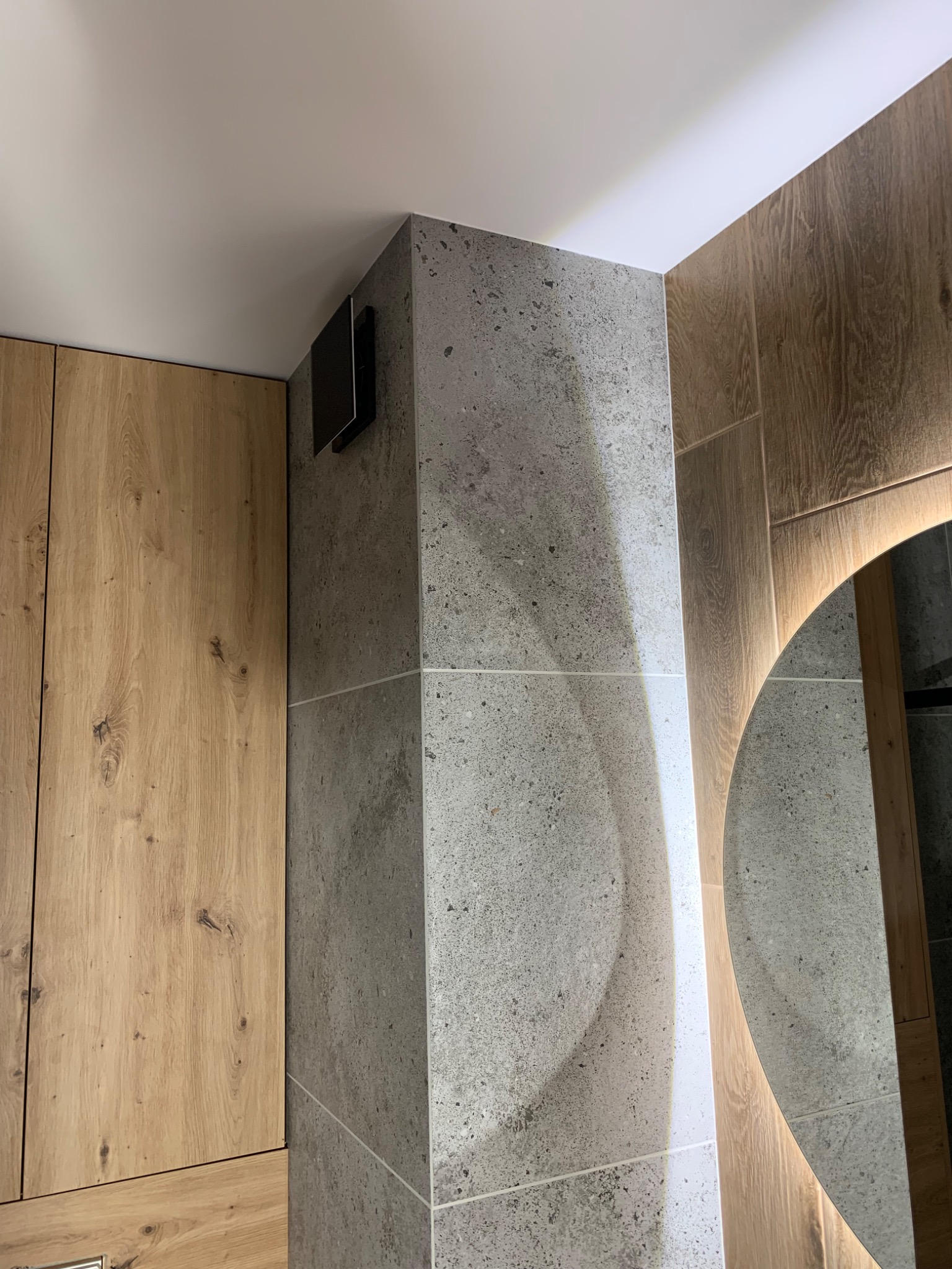 Fragment wykończonej łazienki z drewnianą szafką, szarą ścianą z płytek imitujących kamień oraz okrągłym lustrem z podświetleniem LED, widoczny czarny wentylator na ścianie.