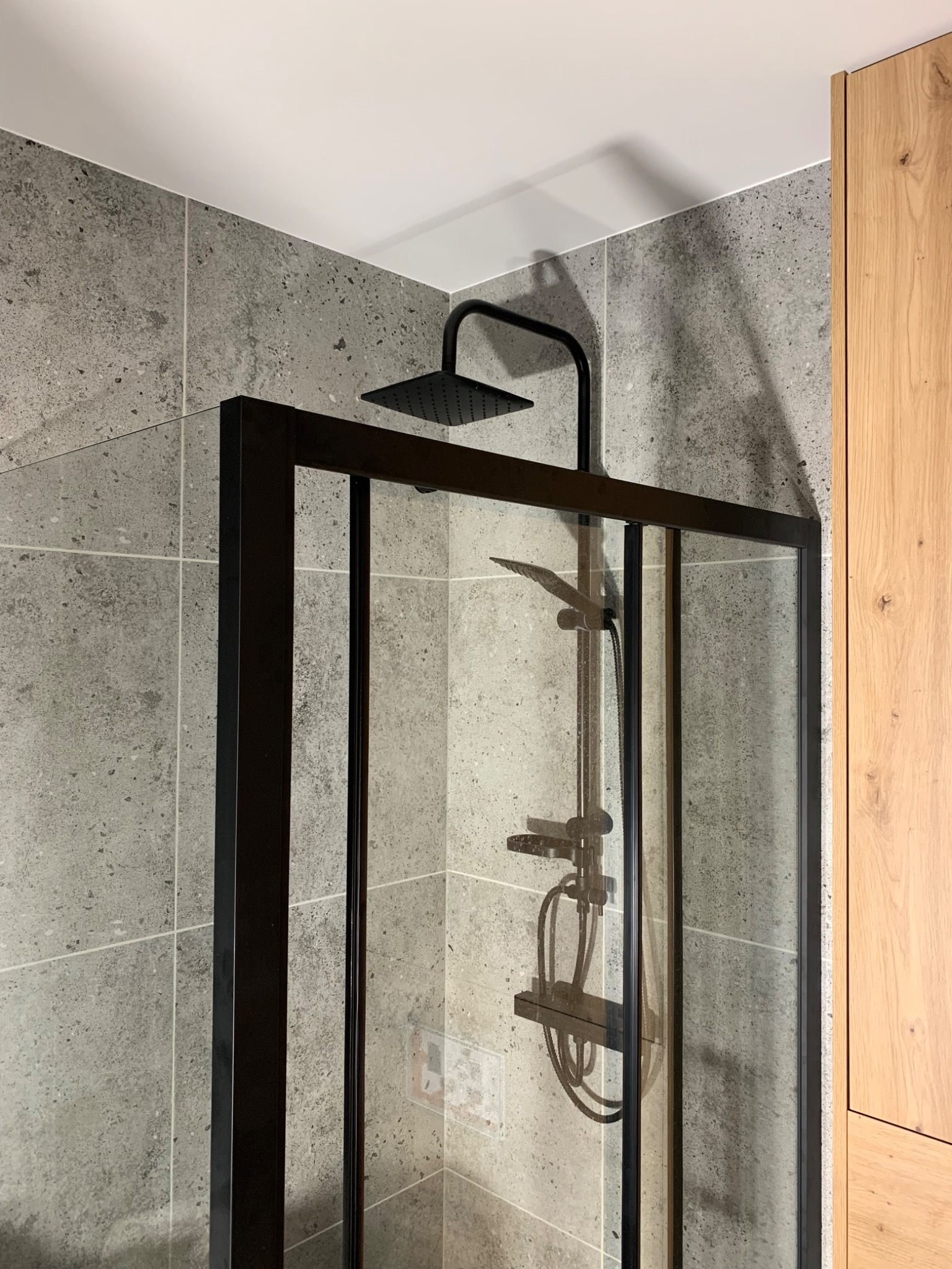 Nowoczesna kabina prysznicowa z czarną armaturą i szarymi płytkami imitującymi kamień, sąsiadująca z drewnianą zabudową meblową.
