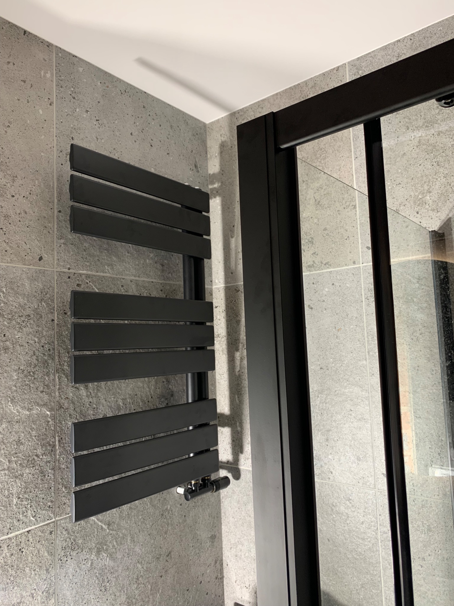 Czarny grzejnik drabinkowy na szarej, kwadratowej ścianie z płytek, obok czarnej ramy kabiny prysznicowej z przezroczystym szkłem.