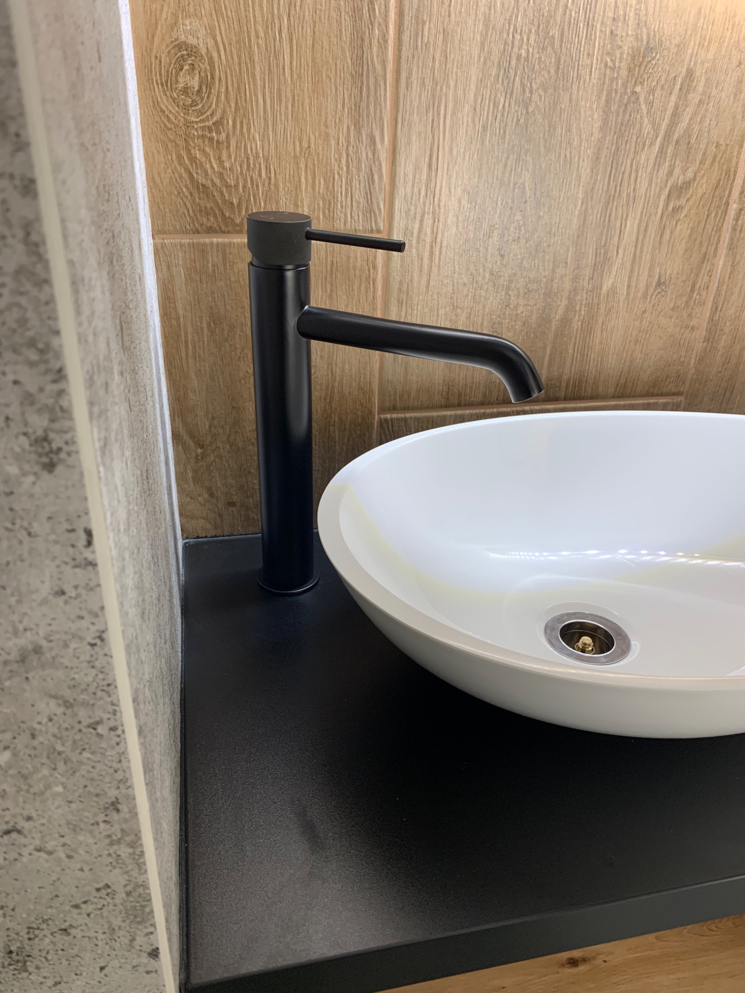 Biała, owalna umywalka nablatowa z czarną, minimalistyczną baterią na czarnym blacie, na tle płytek imitujących drewno i beton