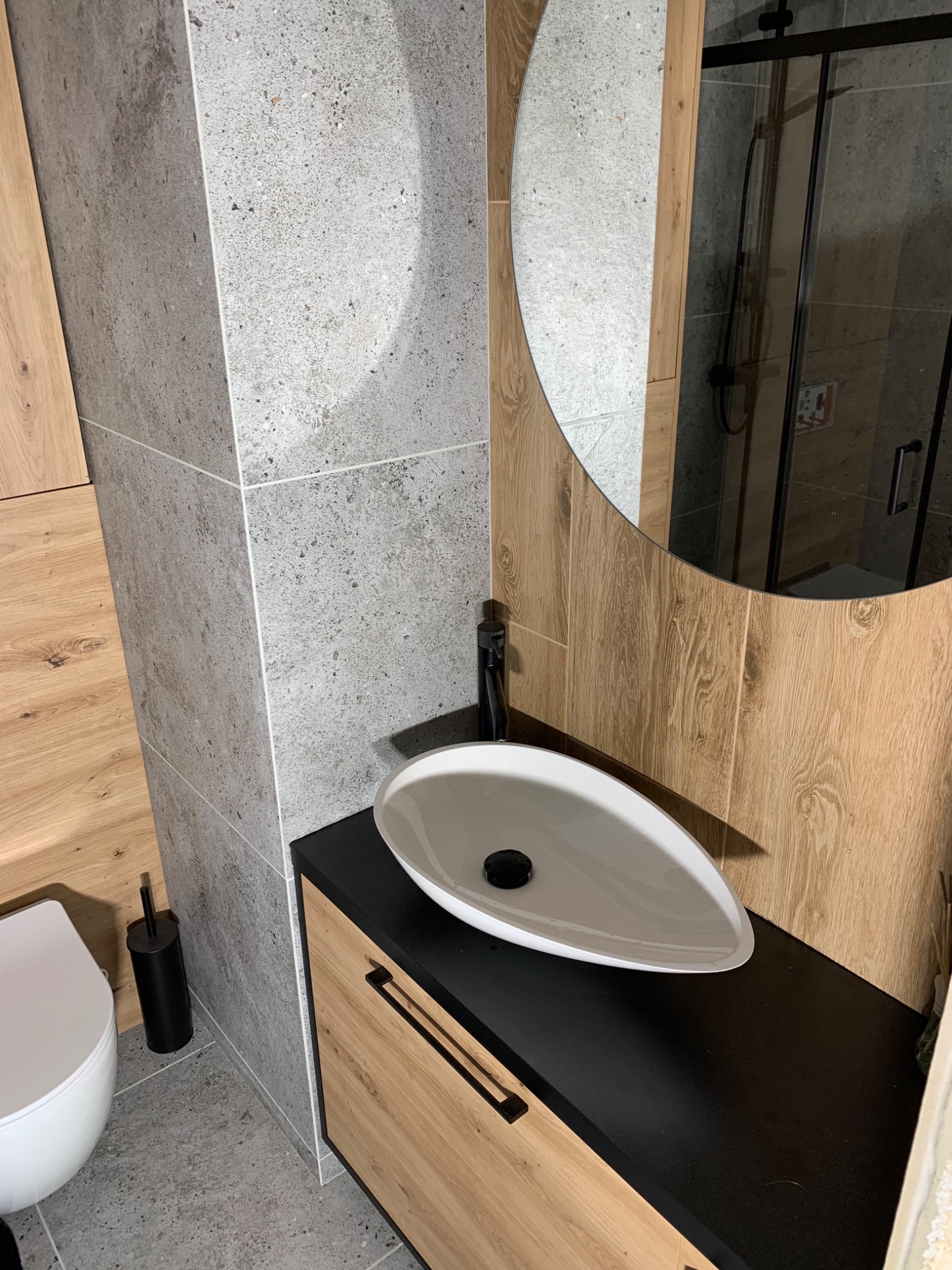 Nowoczesna łazienka z umywalką nablatową w kształcie łzy, czarną armaturą, lustrem w drewnianej ramie i szarą płytką imitującą kamień.