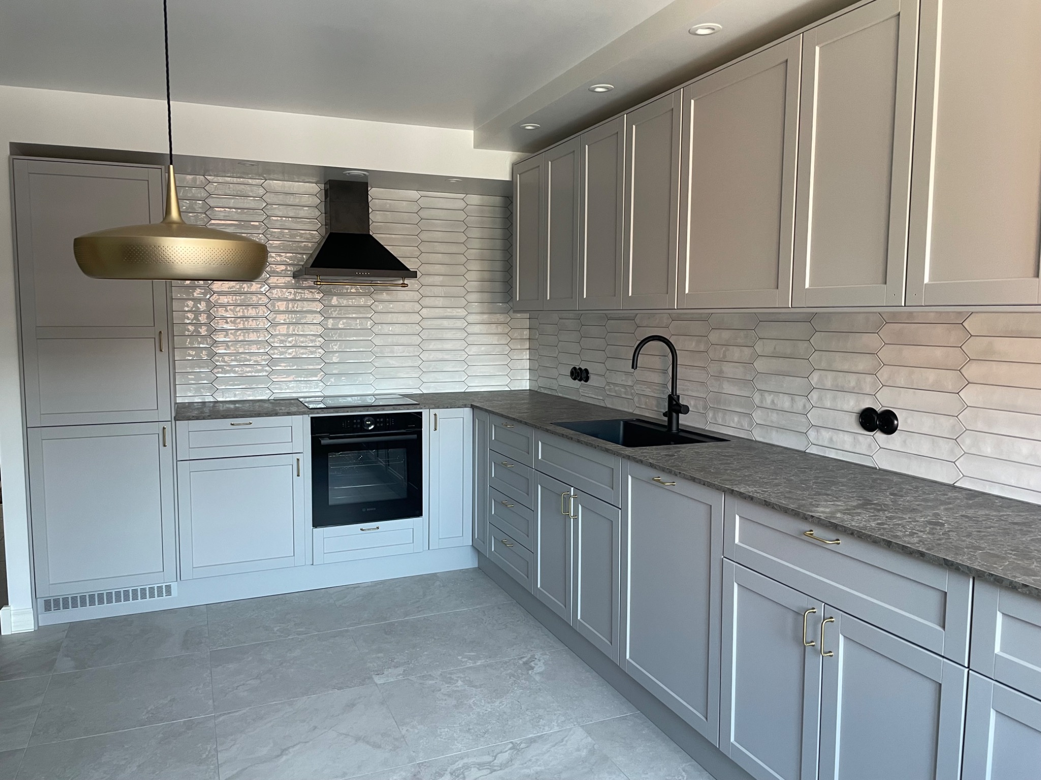 Wykończona kuchnia w odcieniach szarości z płytkami w kształcie wydłużonych rombów na ścianie, czarną armaturą i złotą lampą wiszącą nad blatem.