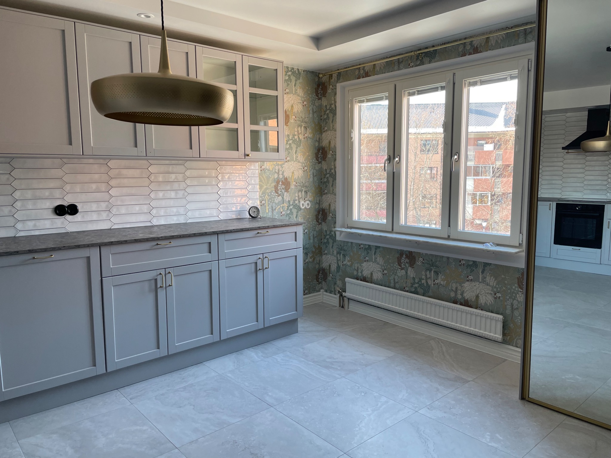 Jasna kuchnia po remoncie z szarymi szafkami, wzorzystą tapetą i heksagonalnymi płytkami na ścianie, oświetlona złotą lampą wiszącą.