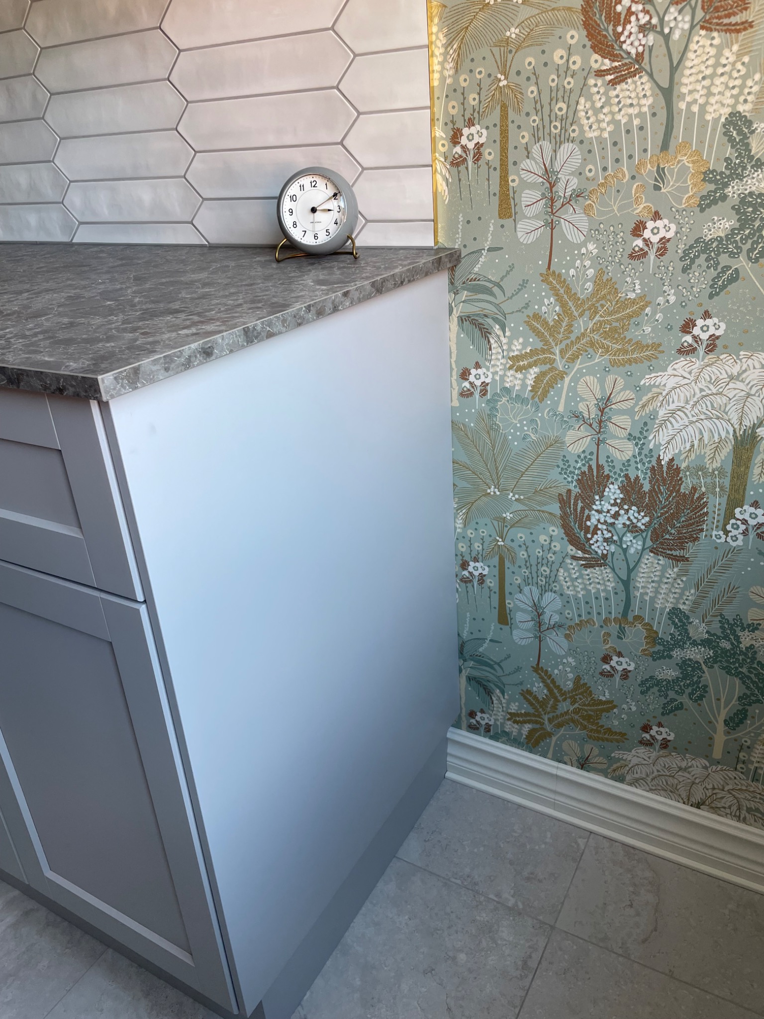 Szara szafka kuchenna z marmurowym blatem, ozdobiona analogowym zegarem, na tle wzorzystej tapety z motywem roślinnym i jasnoszarych, podłużnych płytek ściennych.