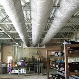 Wnętrze hali produkcyjnej z trzema dużymi, elastycznymi przewodami wentylacyjnymi podwieszonymi do metalowej konstrukcji stropu, widoczne regały z formami oraz pracownicy w tle.