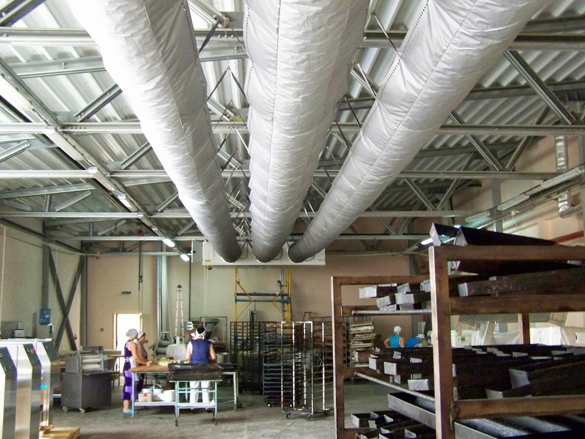 Wnętrze hali produkcyjnej z trzema dużymi, elastycznymi przewodami wentylacyjnymi podwieszonymi do metalowej konstrukcji stropu, widoczne regały z formami oraz pracownicy w tle.