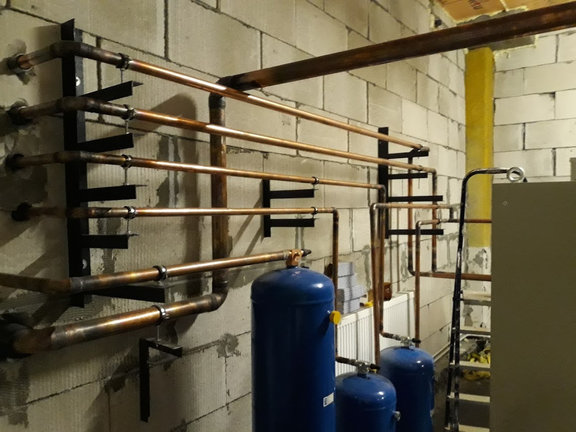 Instalacja z miedzianych rur zamontowana na ścianie z betonowych bloków, widoczne czarne wsporniki, niebieskie zbiorniki ciśnieniowe, drabina i grzejnik.
