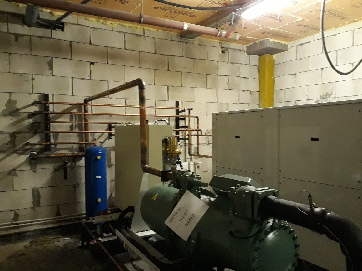 Kompresor chłodniczy z instalacją rurową w surowym pomieszczeniu z bloków betonowych, widoczny niebieski zbiornik ciśnieniowy i tabliczka ostrzegawcza 'WARNING! VALVES CLOSED'.