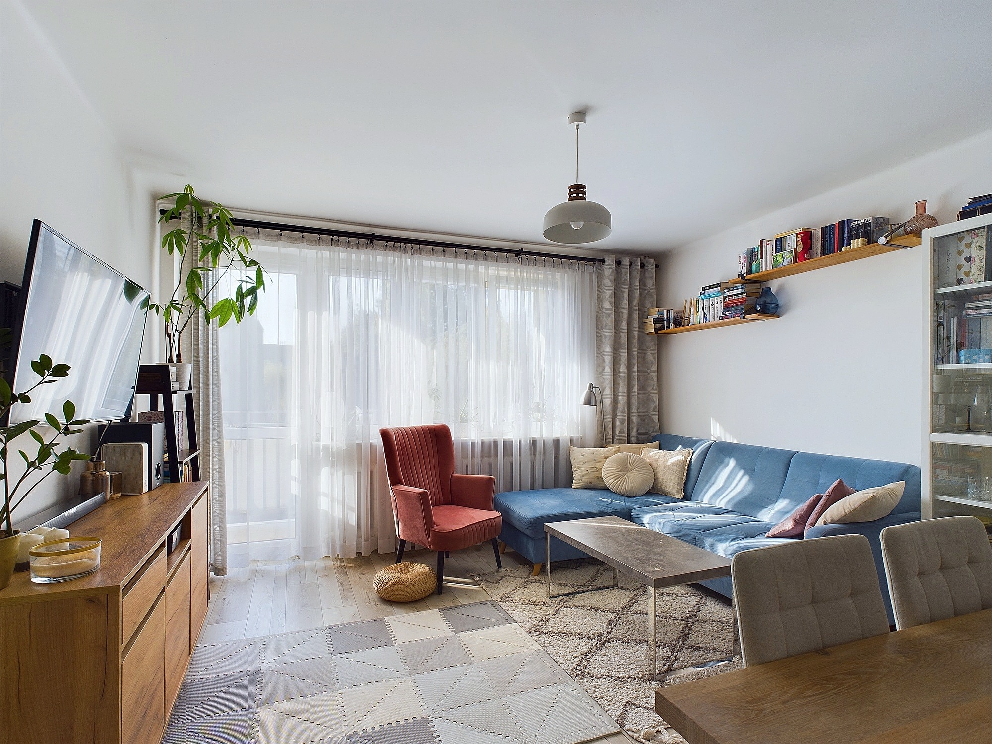 Jasne, przestronne wnętrze salonu z niebieską sofą narożną, ceglastym fotelem, drewnianą komodą z rośliną doniczkową i półką z książkami na ścianie.