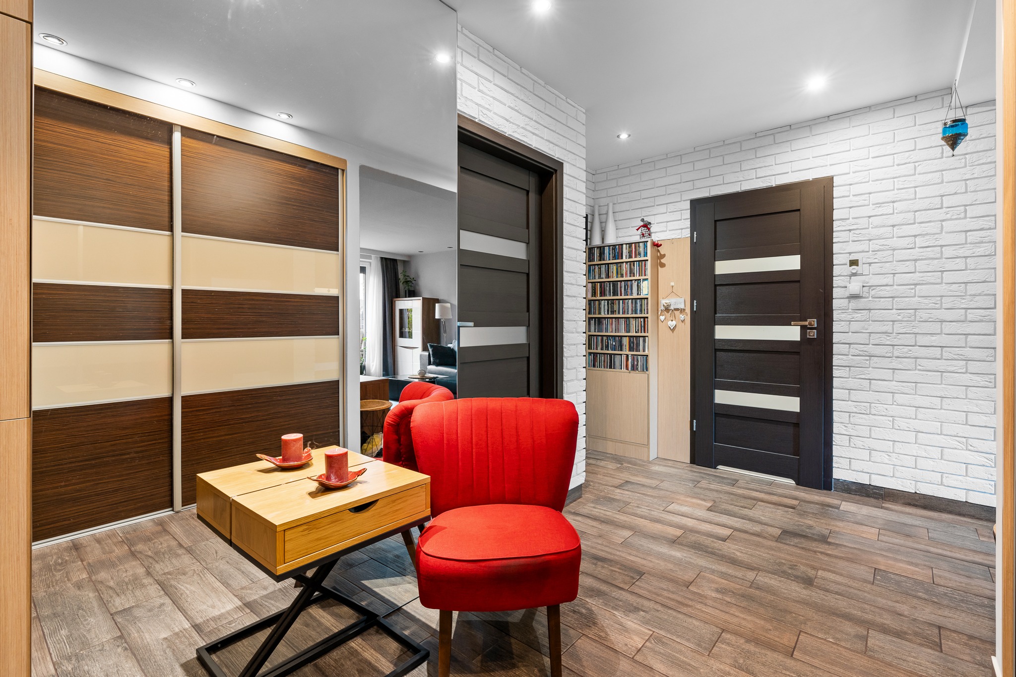 Wnętrze mieszkania z przesuwną szafą w odcieniach brązu i beżu, czerwonym fotelem na drewnianej podłodze, małym drewnianym stolikiem z dwoma świeczkami, białymi cegłami na ścianie i ciemnymi drzwiami.