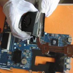 Czyszczenie radiatora i wentylatora procesora laptopa z kurzu przez technika w białych rękawiczkach, widoczna płyta główna, śrubokręt i pomarańczowe tło.