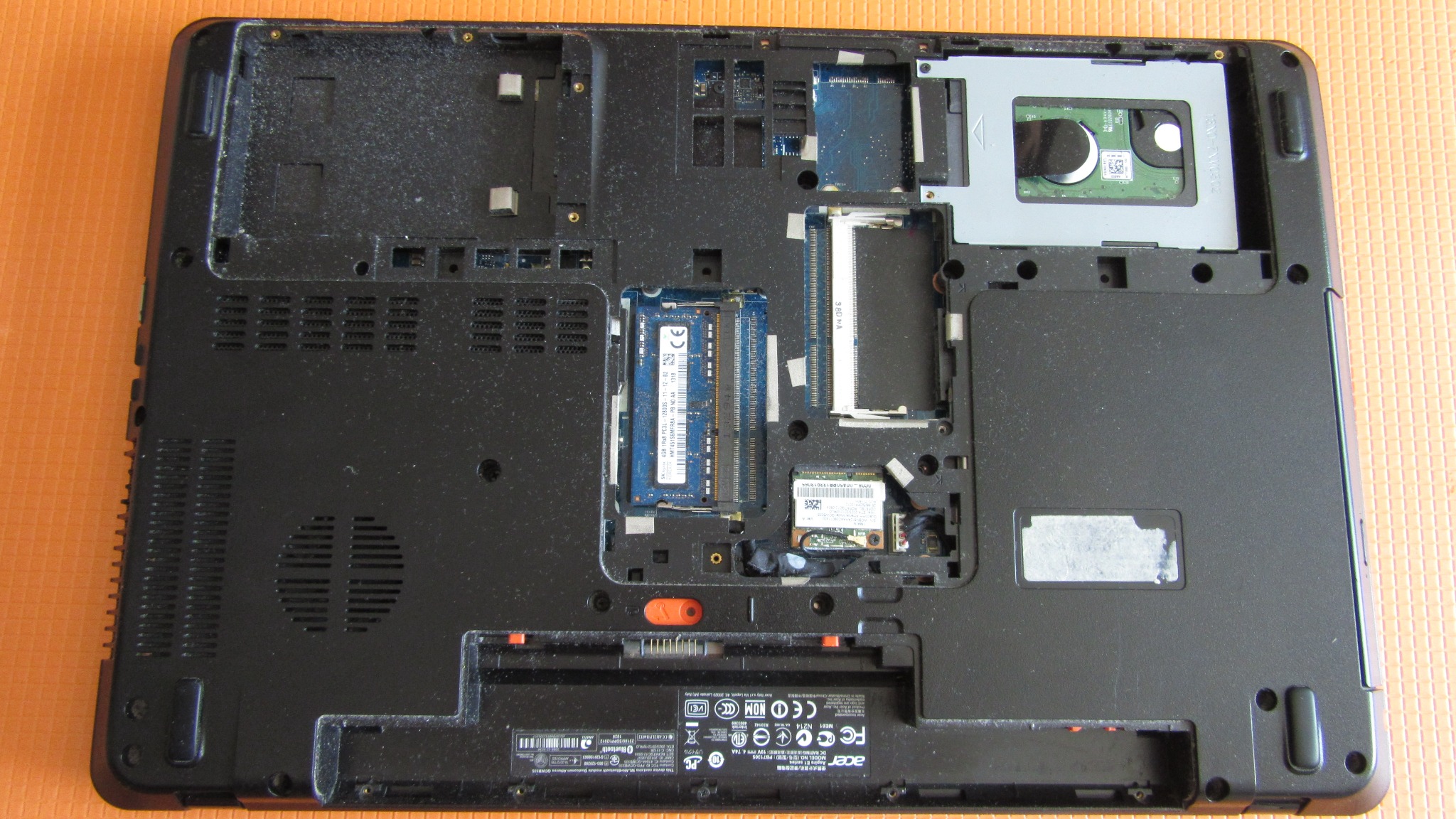 Wnętrze rozebranego laptopa Acer z widocznymi modułami RAM, dyskiem twardym i baterią, leżącego na pomarańczowym tle.