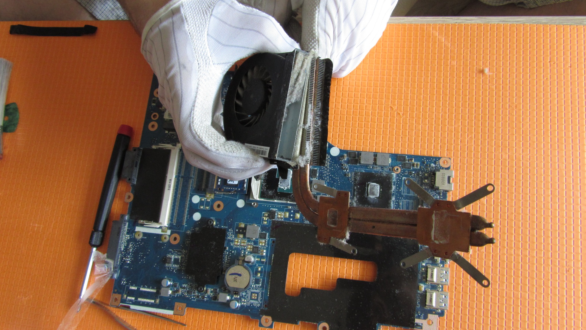 Czyszczenie radiatora i wentylatora procesora laptopa z kurzu przez technika w białych rękawiczkach, widoczna płyta główna, śrubokręt i pomarańczowe tło.
