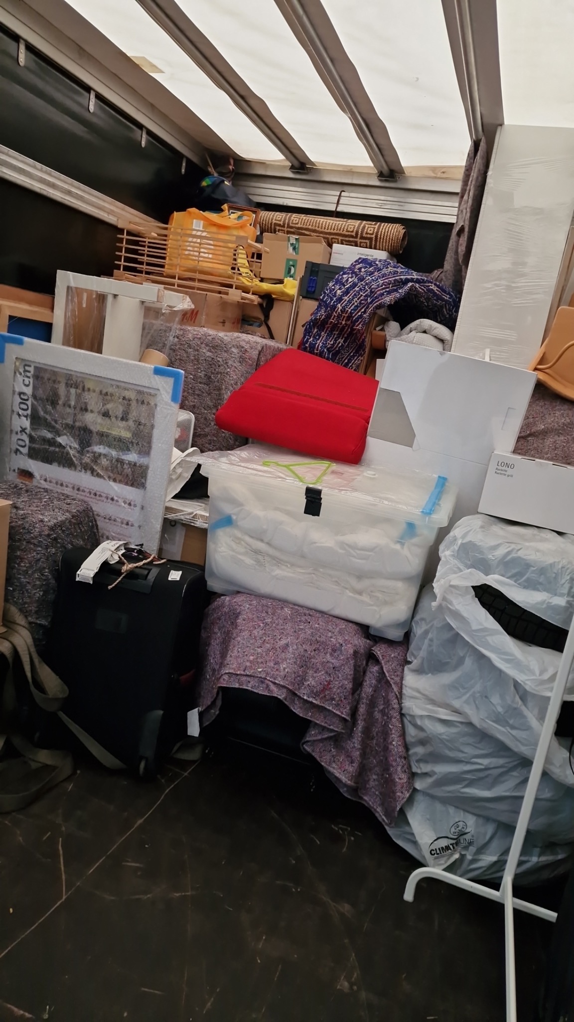 Wnętrze przestrzeni ładunkowej wypełnionej spakowanymi przedmiotami: meblami, obrazem w ramie z napisem '70x100 cm', walizką na kółkach, pudełkami, torbami foliowymi i kocami zabezpieczającymi.