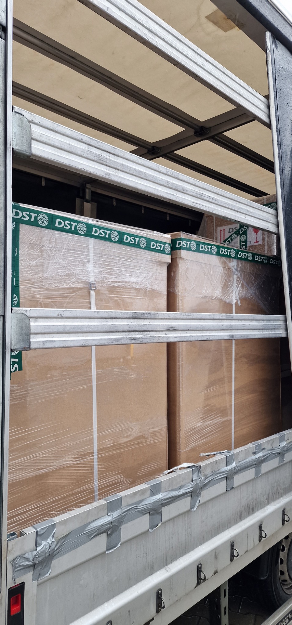 Kartonowe paczki owinięte folią i taśmą z logo DST, umieszczone wewnątrz skrzyni ładunkowej samochodu dostawczego, zabezpieczone metalowymi listwami.