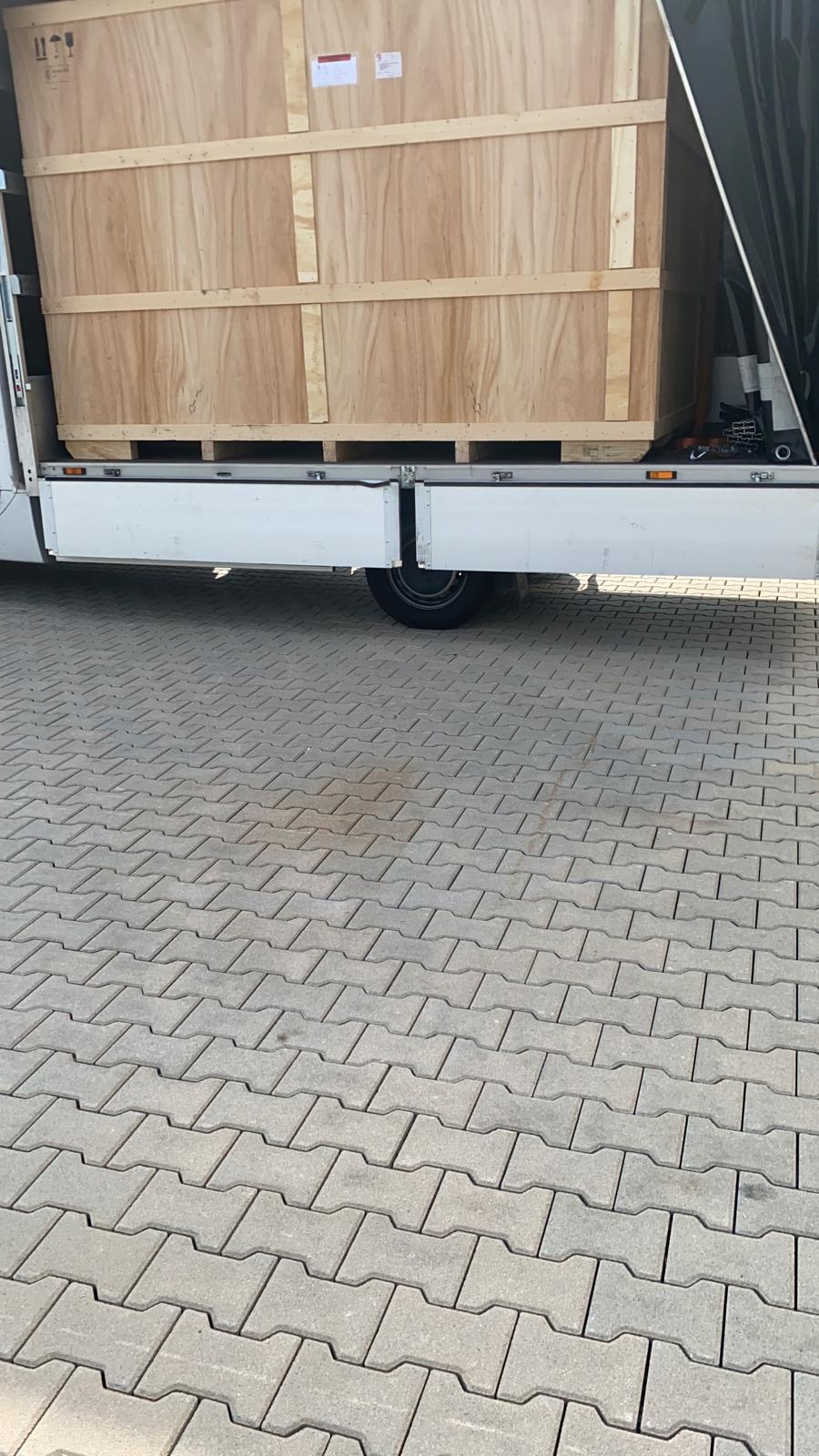 Duża drewniana skrzynia zabezpieczona na palecie, umieszczona w przestrzeni ładunkowej białego pojazdu dostawczego, widoczna na tle kostki brukowej.