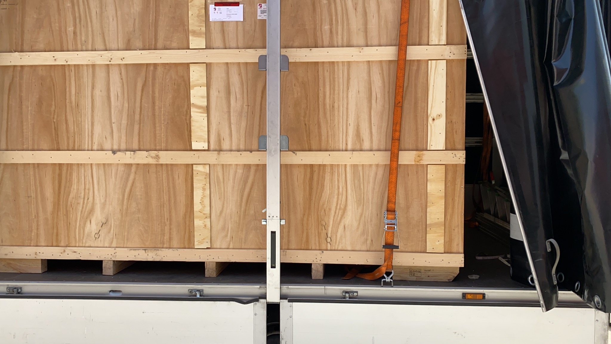 Drewniana skrzynia zabezpieczona pasami w przestrzeni ładunkowej pojazdu, widoczna aluminiowa listwa mocująca i fragment czarnej plandeki.
