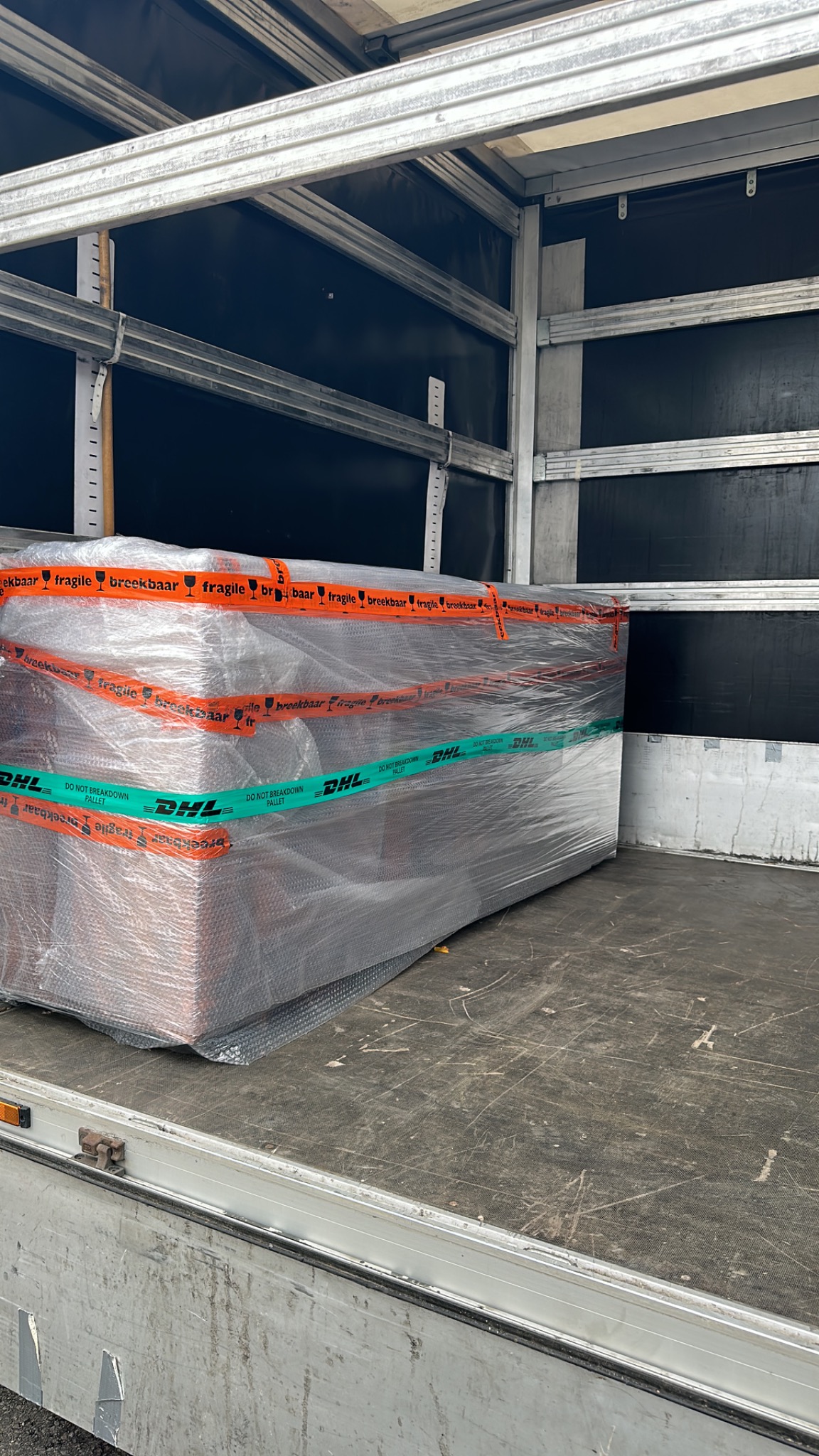 Paczka owinięta folią bąbelkową, zabezpieczona taśmami z napisem 'fragile' i logo DHL, umieszczona w przestrzeni ładunkowej pojazdu.