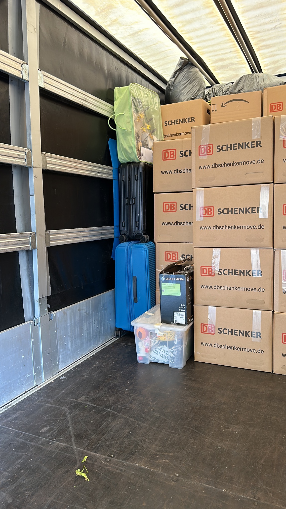 Wnętrze ciężarówki załadowanej kartonami Schenker, walizkami w odcieniach niebieskiego i czerni oraz zieloną torbą, przygotowanymi do transportu podczas przeprowadzki.