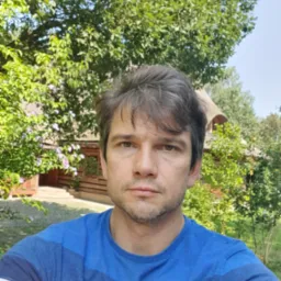 Mężczyzna w niebieskiej koszulce na tle kwitnącego ogrodu i drewnianego domu z gontowym dachem.