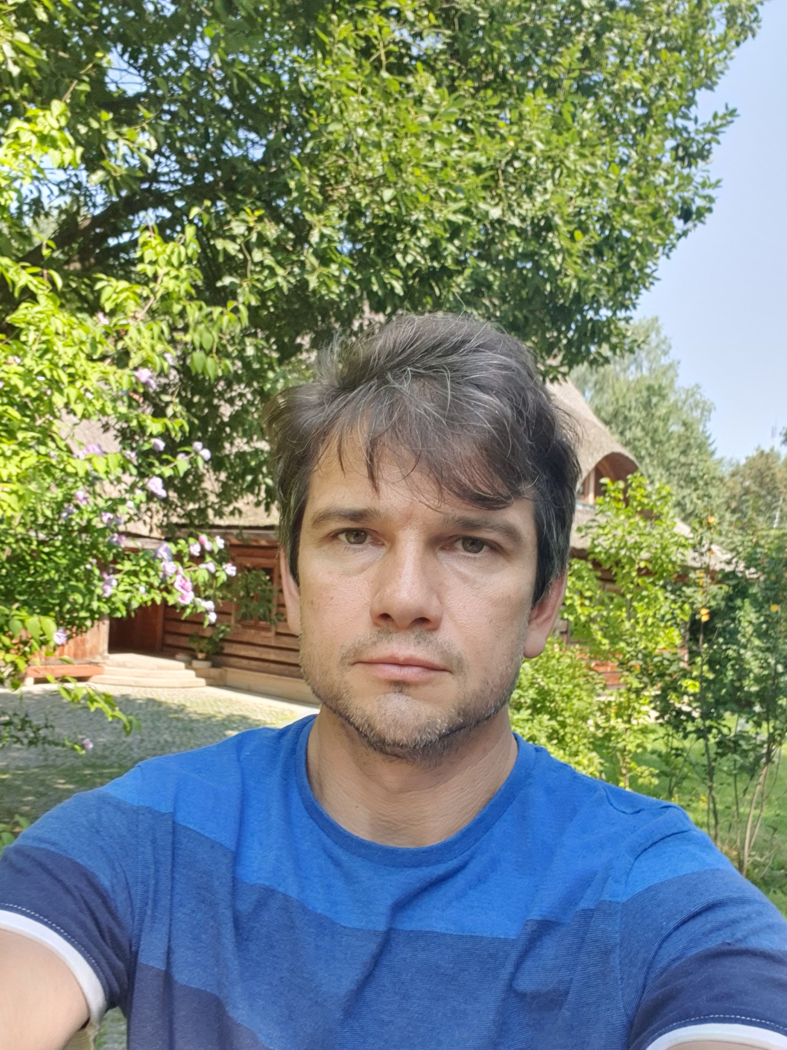 Mężczyzna w niebieskiej koszulce na tle kwitnącego ogrodu i drewnianego domu z gontowym dachem.