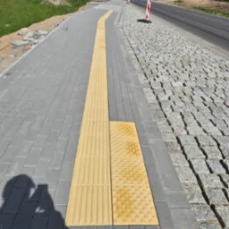 Nowo ułożony chodnik z kostki brukowej z żółtymi płytami dotykowymi dla osób niewidomych, sąsiadujący z drogą wyłożoną kostką granitową, widoczne słupki drogowe.