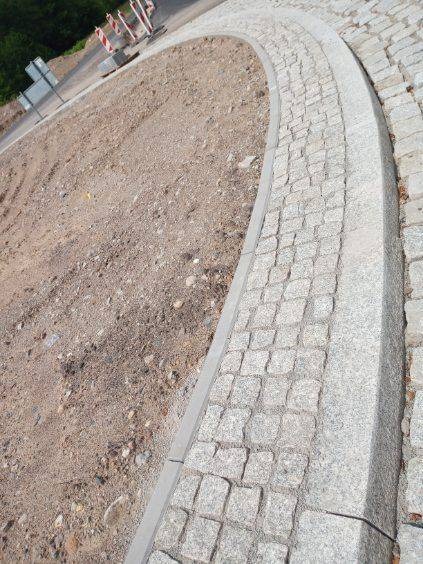 Zakrzywiona ścieżka z kostki granitowej w trakcie budowy, widoczne przygotowane podłoże i krawężniki, pomarańczowe pachołki w tle.