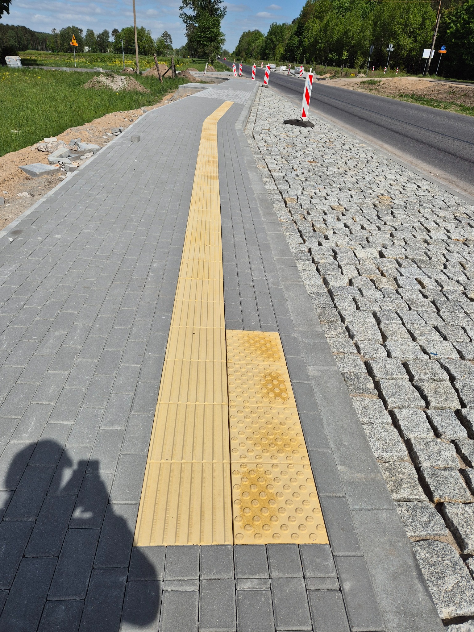 Nowo ułożony chodnik z kostki brukowej z żółtymi płytami dotykowymi dla osób niewidomych, sąsiadujący z drogą wyłożoną kostką granitową, widoczne słupki drogowe.