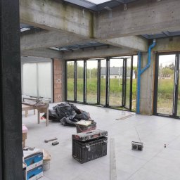 Pal-Mal - Wnętrze w trakcie prac wykończeniowych: widoczne rozłożone składane drzwi okienne z czarnymi ramami, surowy betonowy strop z oknami dachowymi, szare płytki na podłodze, niebieskie rury, narzędzia...