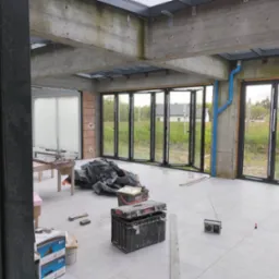Wnętrze w trakcie prac wykończeniowych: widoczne rozłożone składane drzwi okienne z czarnymi ramami, surowy betonowy strop z oknami dachowymi, szare płytki na podłodze, niebieskie rury, narzędzia...