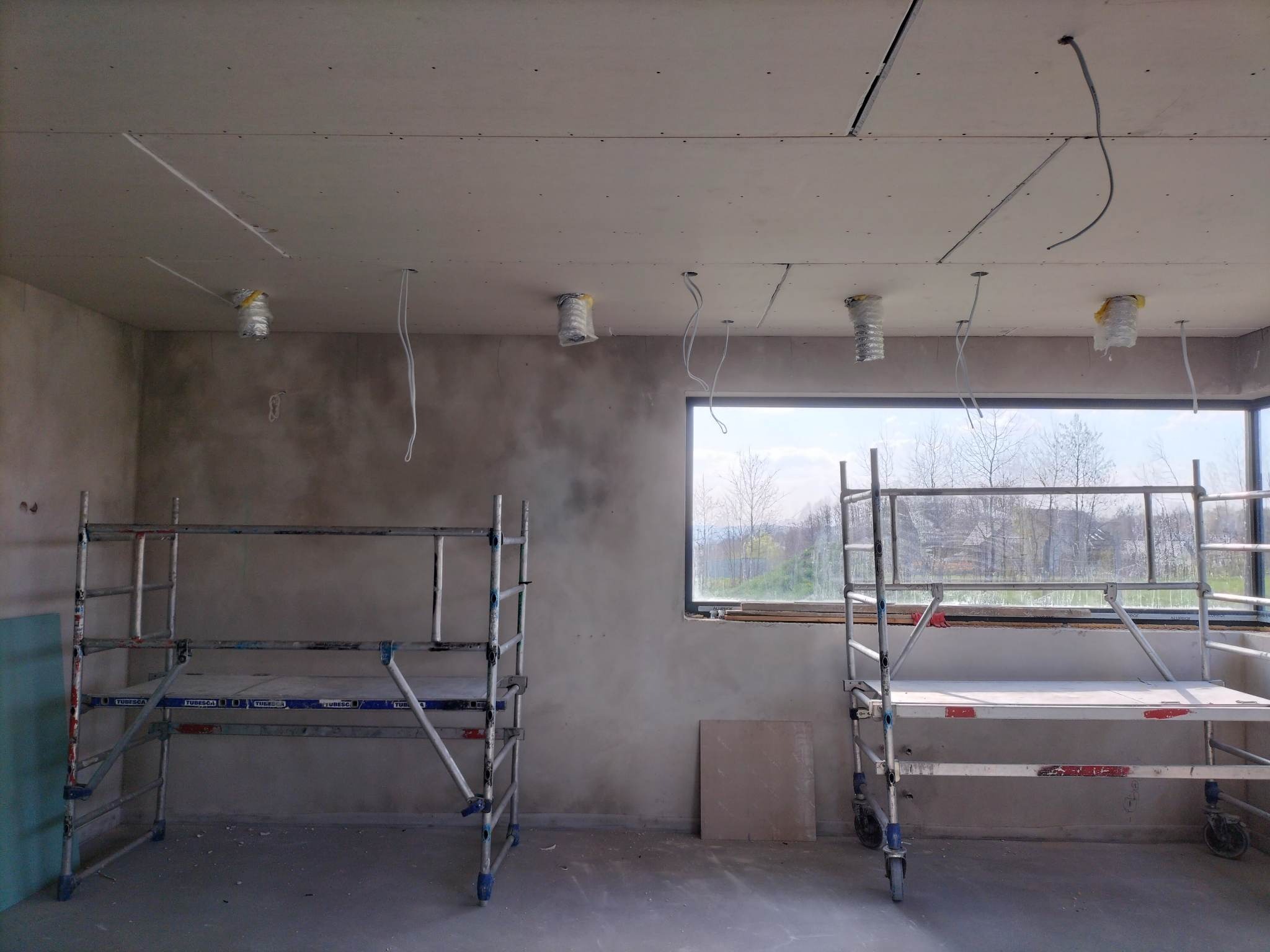 Wnętrze w trakcie prac wykończeniowych: sufit z zamontowaną płytą gipsowo-kartonową z wystającymi przewodami elektrycznymi, widoczne rusztowania i niezabezpieczona ściana w tle.