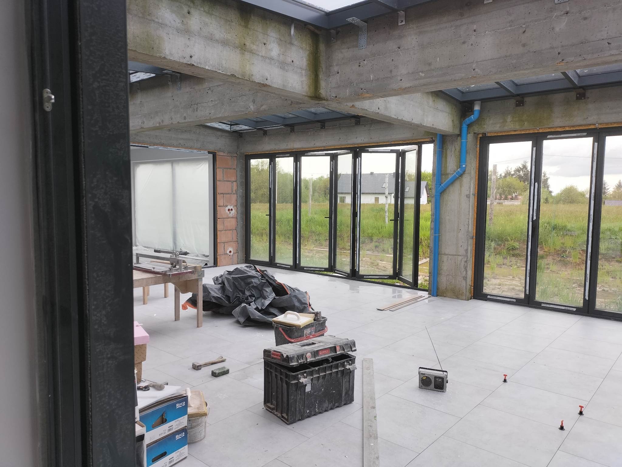 Wnętrze w trakcie prac wykończeniowych: widoczne rozłożone składane drzwi okienne z czarnymi ramami, surowy betonowy strop z oknami dachowymi, szare płytki na podłodze, niebieskie rury, narzędzia...