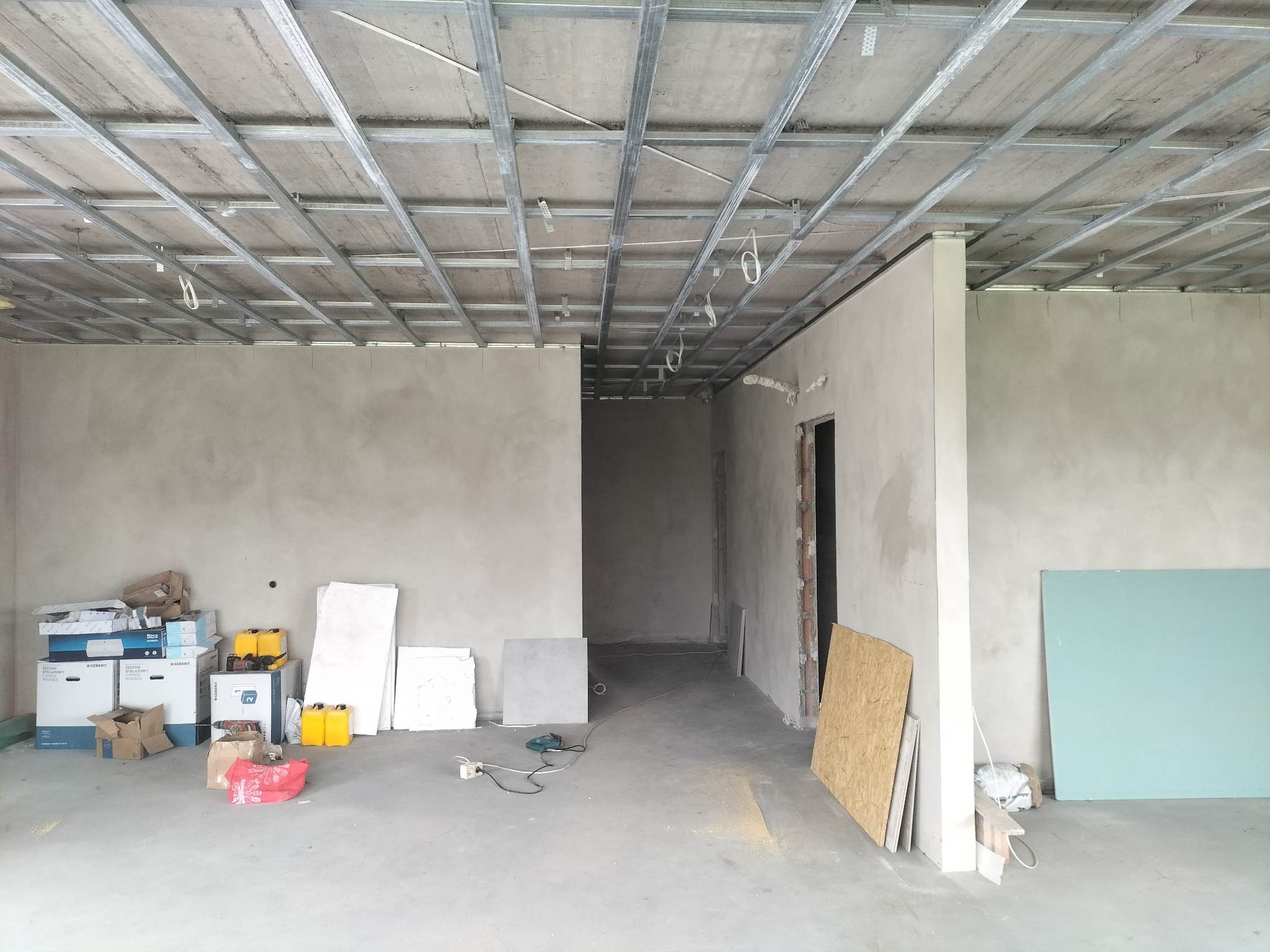 Surowy stan wnętrza w trakcie remontu: widoczne metalowe profile sufitowe, otynkowane ściany, podłoga wylewka betonowa, płyty kartonowo-gipsowe, narzędzia i materiały budowlane, brak wykończenia.