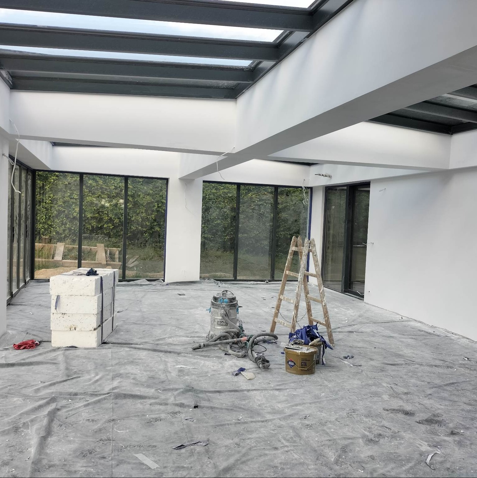 Wnętrze w trakcie prac wykończeniowych: świeżo pomalowane białe ściany, okna z widokiem na zieleń, zabezpieczona folią podłoga, drabina oparta o ścianę, odkurzacz przemysłowy i wiadro z farbą.