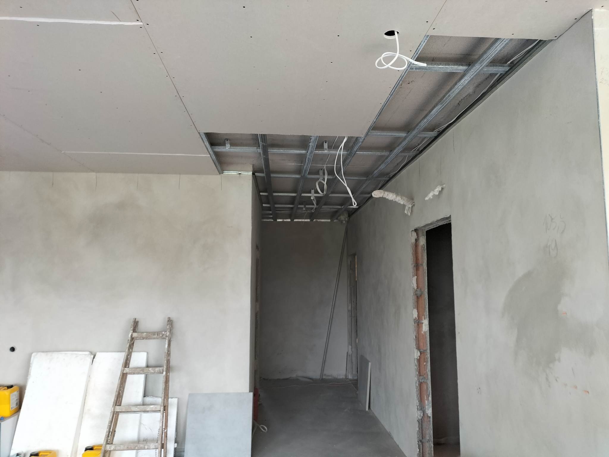 Wnętrze w trakcie wykańczania z widoczną konstrukcją metalową podwieszanego sufitu, częściowo pokrytą płytami gipsowo-kartonowymi, oraz ścianami w stanie surowym, drabina oparta o ścianę, przewody...