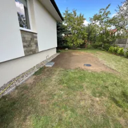 Przygotowany teren pod trawnik z widoczną studzienką kanalizacyjną, obok ściana budynku z kranem i ułożonymi kamieniami ozdobnymi.