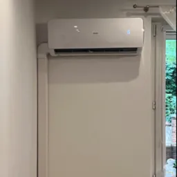 Biała jednostka klimatyzacyjna AUX zamontowana na ścianie w pomieszczeniu, z widocznymi rurami odprowadzającymi i wyświetlaczem temperatury ustawionym na 15 stopni, obok okna z widokiem na zieleń.