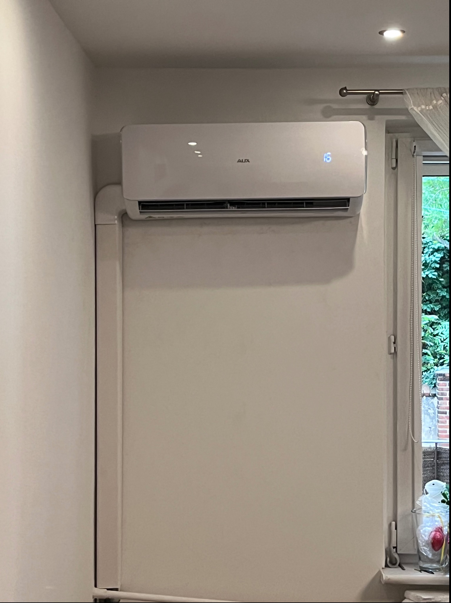 Biała jednostka klimatyzacyjna AUX zamontowana na ścianie w pomieszczeniu, z widocznymi rurami odprowadzającymi i wyświetlaczem temperatury ustawionym na 15 stopni, obok okna z widokiem na zieleń.