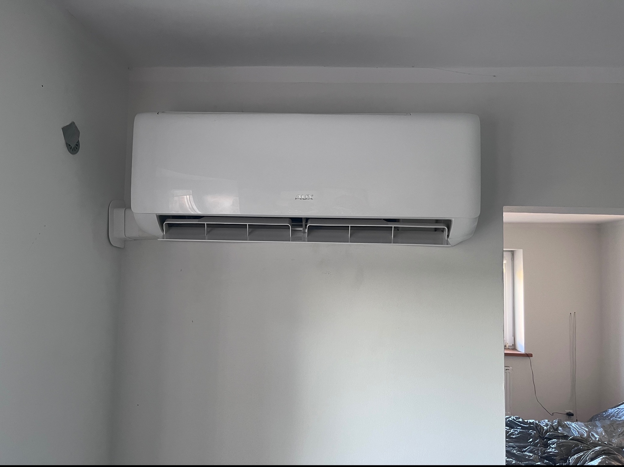 Biały klimatyzator marki AUX zamontowany na białej ścianie w rogu pokoju, widoczne ślady po usuniętej tapecie i przygotowania do malowania.