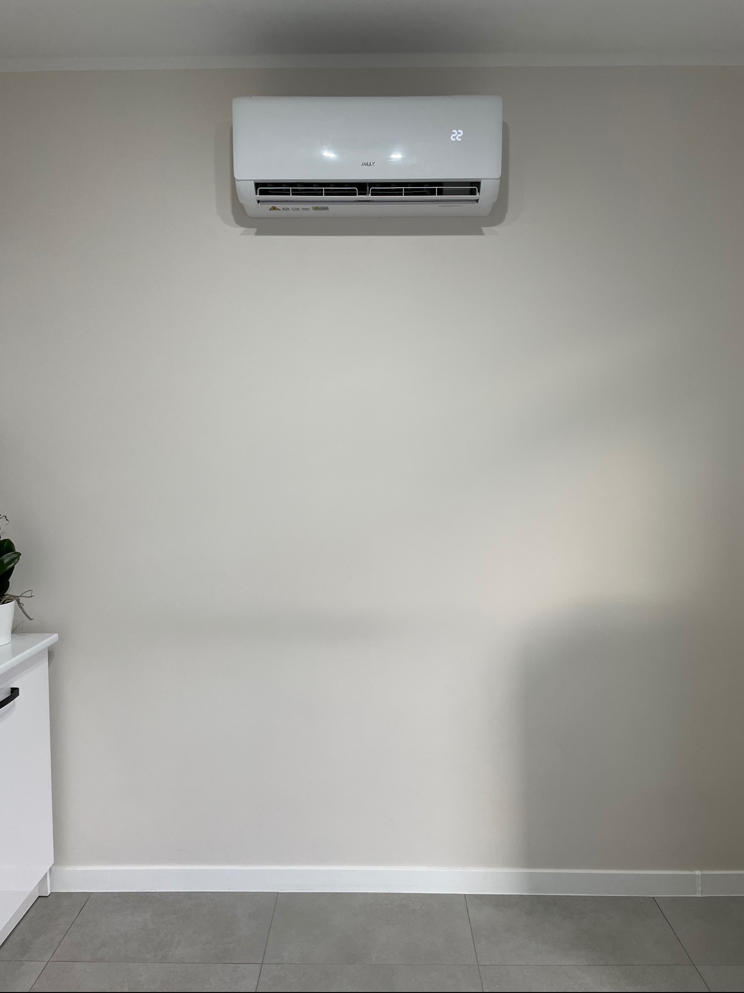 Biała klimatyzacja ścienna z wyświetlaczem temperatury 22 stopnie Celsjusza, zamontowana na jasnej ścianie w minimalistycznym wnętrzu z szarą podłogą i fragmentem białej szafki z rośliną.