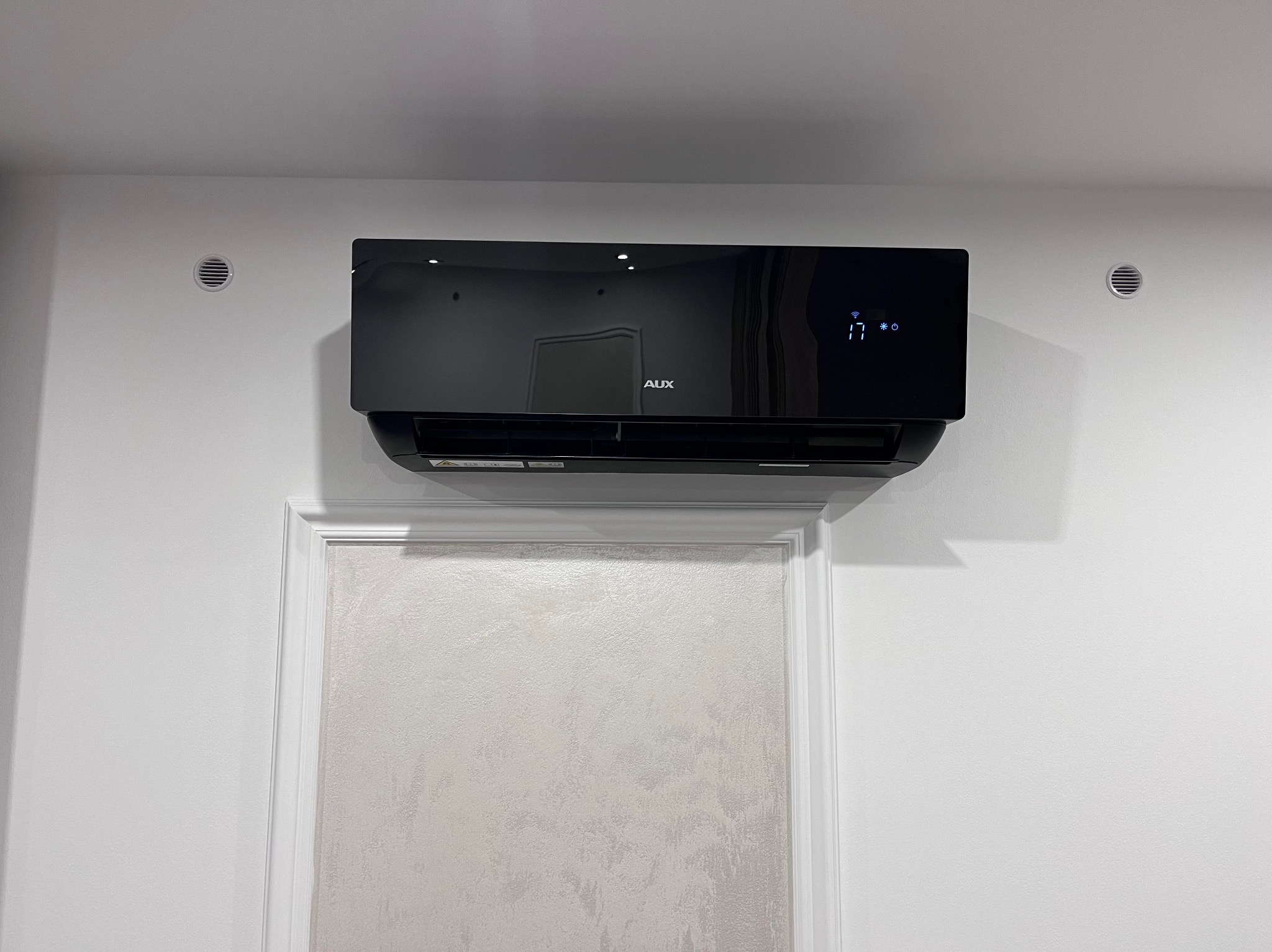 Czarny klimatyzator AUX zamontowany na białej ścianie nad ozdobnym panelem ściennym, widoczne kratki wentylacyjne po bokach.