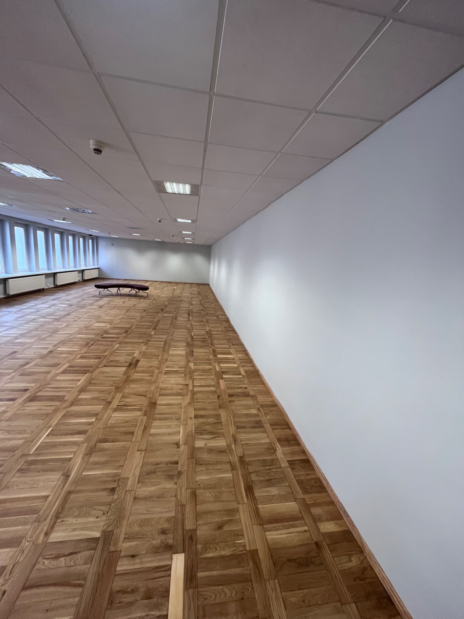 Puste, jasne biuro z parkietem w jodełkę, białymi ścianami i rzędem okien w tle. Na suficie widoczne panele i czujnik dymu.