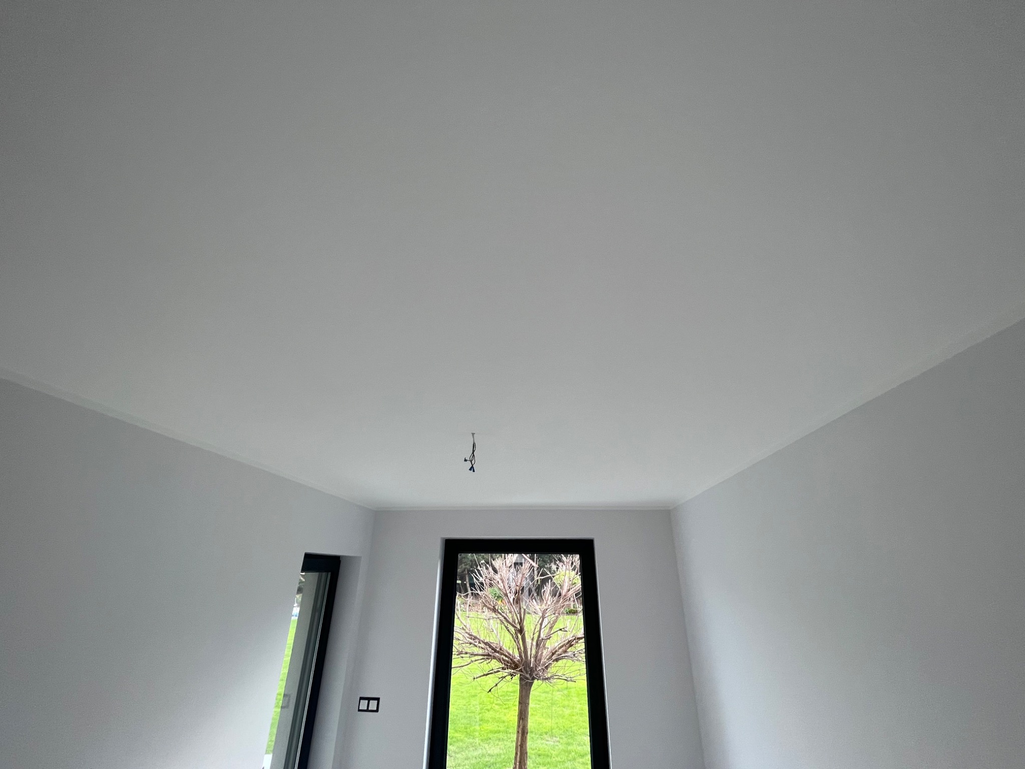 Świeżo pomalowane na biało wnętrze pokoju z widokiem na zielony trawnik przez duże okno. Na suficie widoczne kable od lampy.
