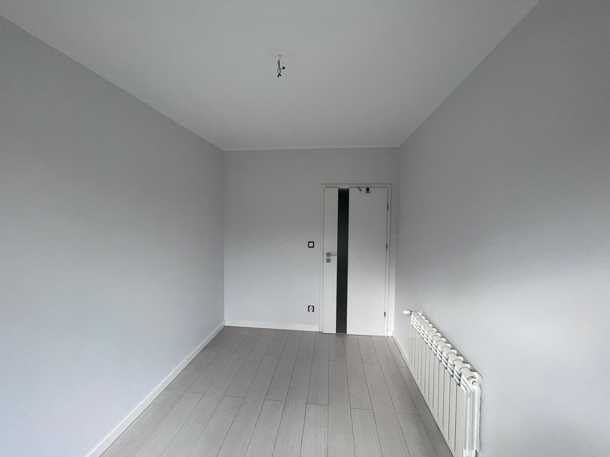 Pomalowane na szaro wnętrze pokoju z panelami podłogowymi, białymi listwami przypodłogowymi, grzejnikiem i drzwiami z czarnym paskiem; widoczne gniazdka elektryczne i wiszące kable od oświetlenia.